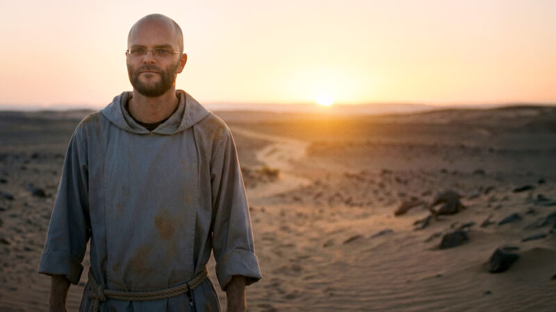 A cinematic still portrait of Fr. Angelus Montgomery, known for the Poco a Poco podcast, wearing a worn grey Franciscan-style habit and rope cincture, walking forward with a calm, hopeful expression in a vast desert landscape. The image captures the powerful moment he concludes his 40-day desert journey, as the warm golden light of a desert sunrise breaks on the horizon, symbolizing how to return with renewed faith and quiet conviction.