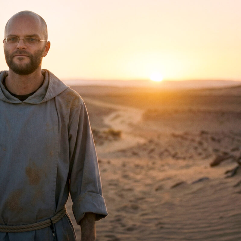 A cinematic still portrait of Fr. Angelus Montgomery, known for the Poco a Poco podcast, wearing a worn grey Franciscan-style habit and rope cincture, walking forward with a calm, hopeful expression in a vast desert landscape. The image captures the powerful moment he concludes his 40-day desert journey, as the warm golden light of a desert sunrise breaks on the horizon, symbolizing how to return with renewed faith and quiet conviction.